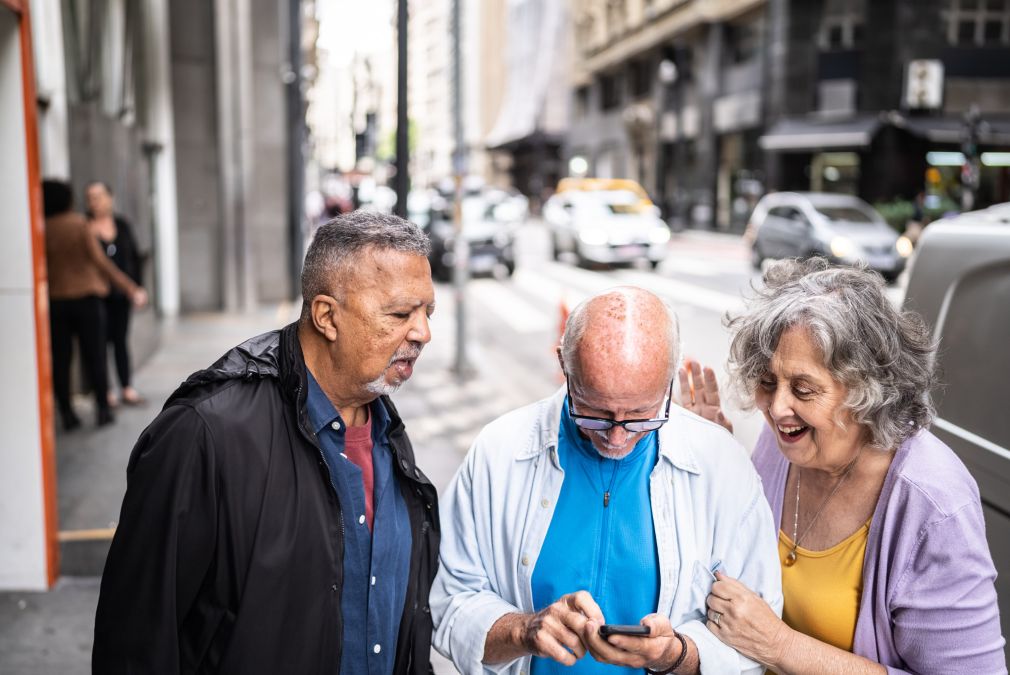 Trois personnes âgées souriantes regardant et utilisant un smartphone ensemble sur un trottoir en ville